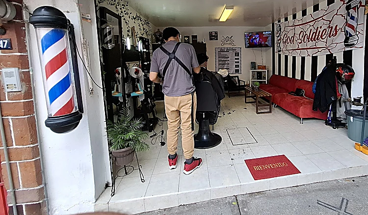 Entrada del local desde la calle con la lona God Soldiers arriba, el poste de barbero y el sillón rojo al fondo.