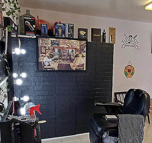 Espejo con focos redondos junto a la pared de ladrillo negro, con máquinas de corte en la repisa y una silla de barbería