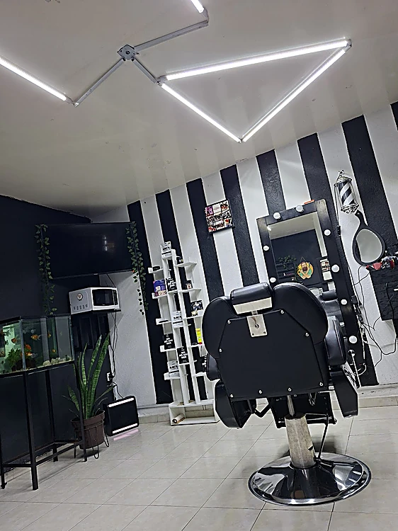 Silla de barbería negra frente al muro de franjas negras y blancas, bajo las barras de luz LED del techo y junto al espejo de focos.