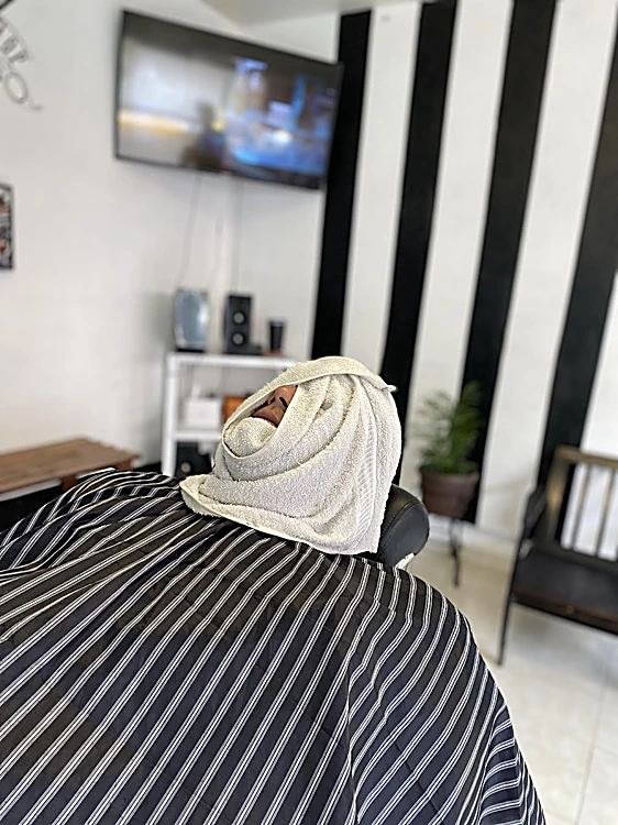 Cliente con el rostro envuelto en una toalla blanca y capa de barbero a rayas.