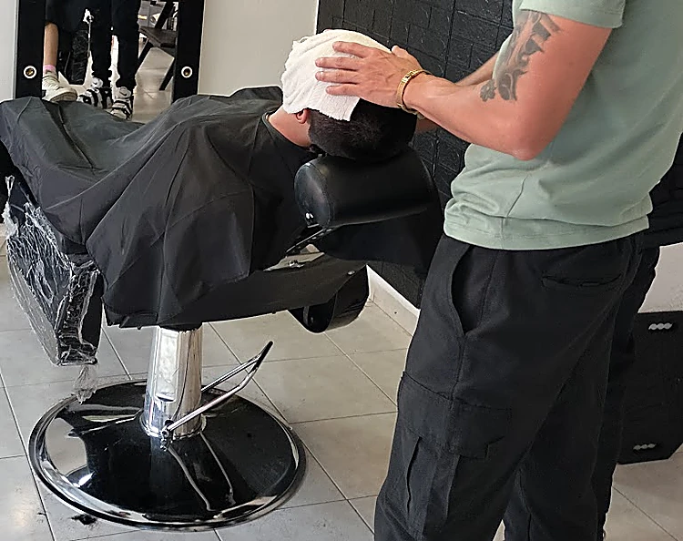 Barbero con brazo tatuado sostiene una toalla caliente sobre la cabeza de un cliente reclinado en la silla.