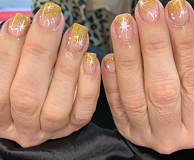 Dos manos con uñas cortas en tono nude, puntas de glitter dorado y estrellas blancas pintadas.